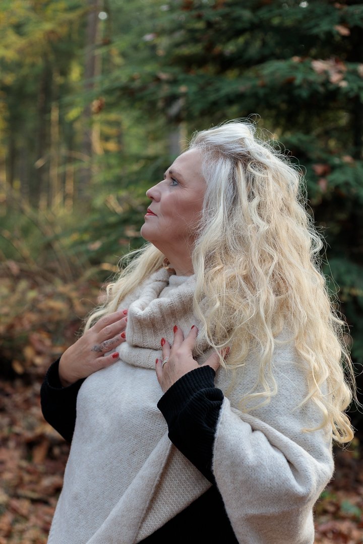 Een vrouw met lang, golvend blond haar staat in een bos, gekleed in een beige, wollen poncho, met haar handen op haar borst, kijkend omhoog, dromerige fine art foto.