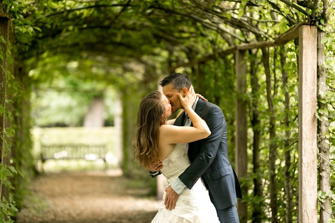 Een bruidspaar kust elkaar liefdevol onder groene, natuurlijk bogen van een kasteeltuin in Loosdrecht