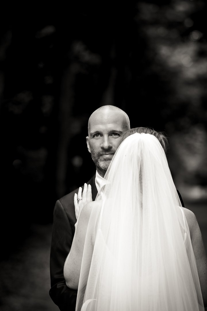 Klassieke zwart-wit foto van een bruid en een bruidegom. De bruid draagt een witte jurk met lange sluier, en de bruidegom draagt een zwart pak. Bruidegom kijkt indringend in de camera tijdens wedding shoot
