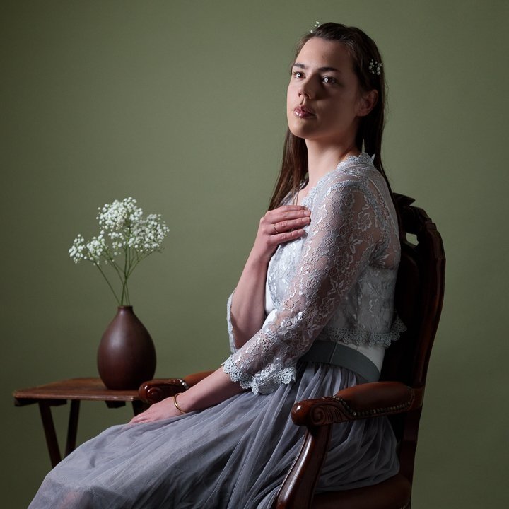 Een jonge vrouw zit op een houten stoel, draagt een wit kanten blouse en een grijze rok, met bloemetjes in haar haren, tegen een groen achtergrond, naast een tafel met een bruine vaas met witte bloemen.