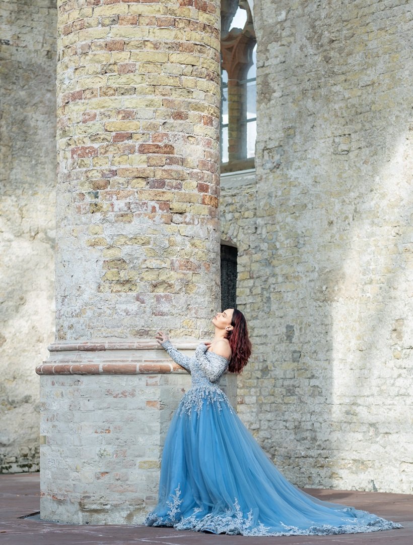 Vrouw in een blauwe jurk met zilveren details leunt tegen een grote stenen muur van een oude kerk tijdens een fotosessie