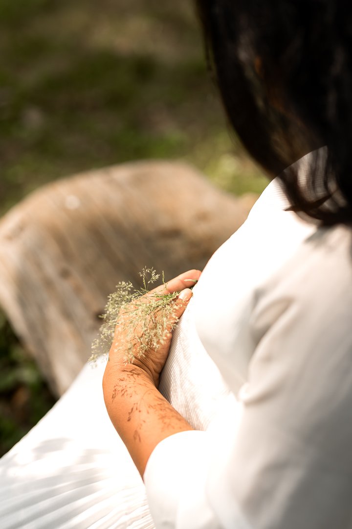 Serene foto, een close-up van een vrouw met zwart haar, die een takje met kleine bloemen vasthoudt, dit brengt mooie schaduw op haar arm. Ze zit op een boomstam, met een witte kleding, zwangerschapsfotografie