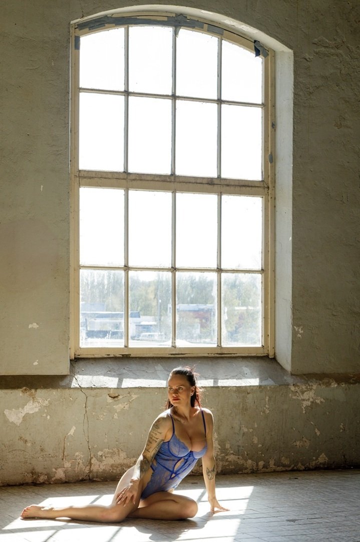 Een vrouw in blauwe lingerie zit op de vloer voor een grote, oude fabriekraam, met zonlicht dat naar binnen schijnt.