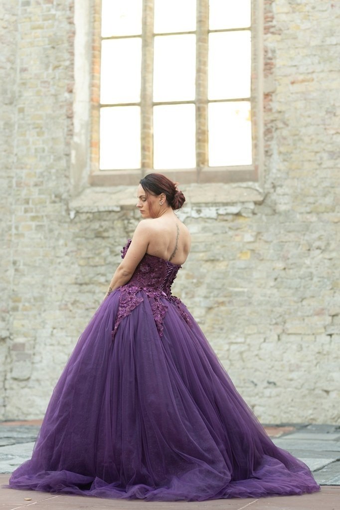 Vrouw in een paarse baljurk met strapless top, staande voor een oude stenen muur met een groot, oud raam op de achtergrond in een kerk met veel natuurlijk licht.