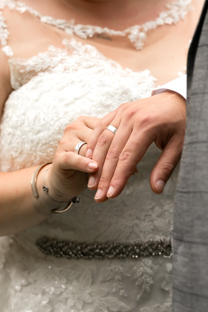 Close-up van een getrouwd stel met hun handen en trouwringen, de bruid draagt een witte kanten trouwjurk en sieraden, de bruidegom draagt een grijze pak. Ze houden elkaars handen vast.