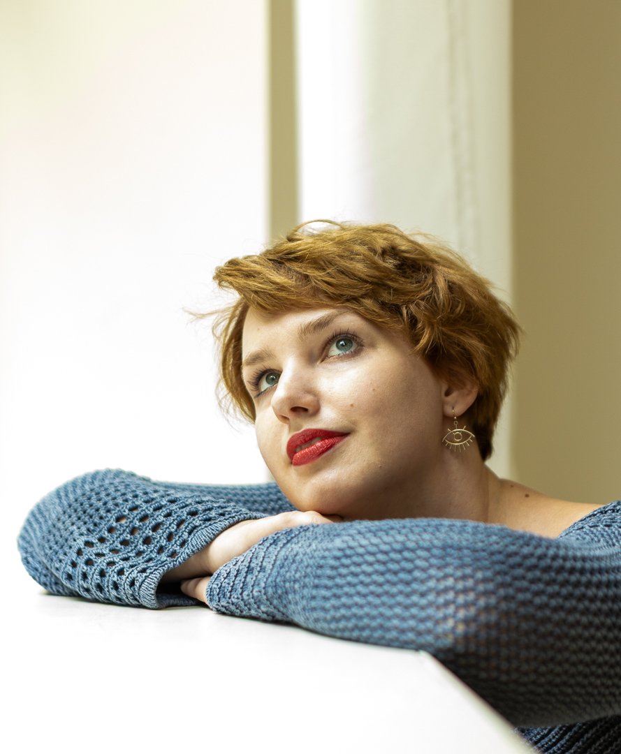 Een vrouw met kort, kastanjebruin haar, haar hoofd rust op haar armen, gekleed in een blauw gehaakt truitje, met een lichte achtergrond. Klassieke look op de foto