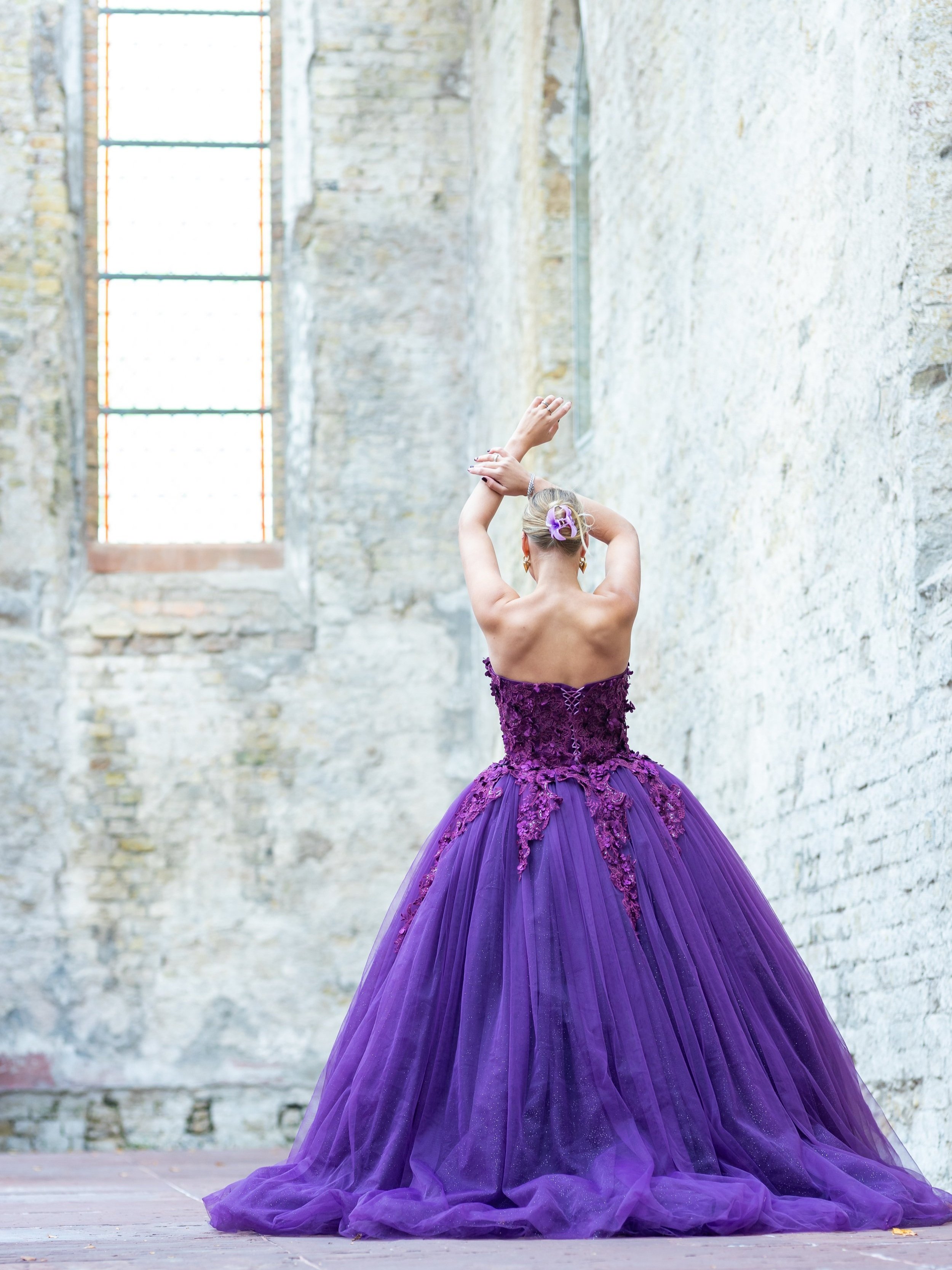 Een vrouw in een strapless paarse galajurk met een hoepel rok, blond opgestoken haar. Zij staat in een oude kerk, met witte bakstenen muren en een raam. Ze draait zich om met haar handen boven haar hoofd, bijna kunst