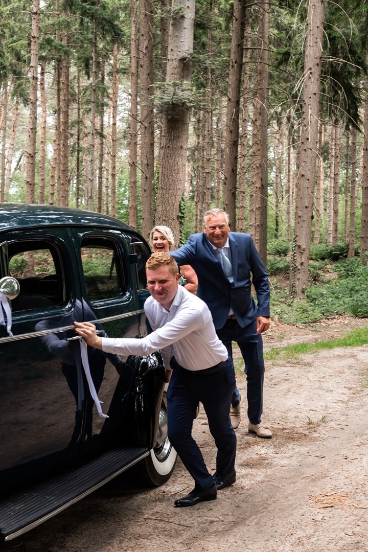Bruidspaar en chauffeur die lachen en plezier maken naast een klassieke zwarte auto in een bos, zij duwen de auto omdat deze niet start, tijdens een bruiloft
