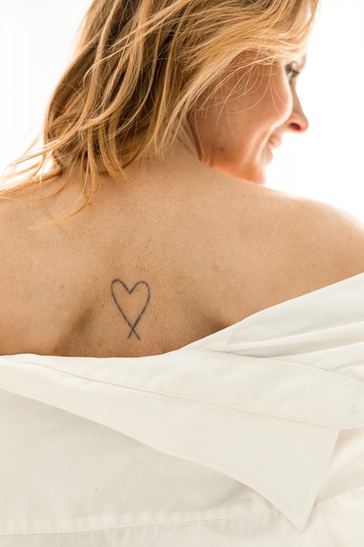 Een vrouw met koperkleurig haar staat in een blouse,die is afgezakt en daardoor zie je hartvormige tatoeage tussen haar schouderbladen. Boudoirfotografie in Drenthe