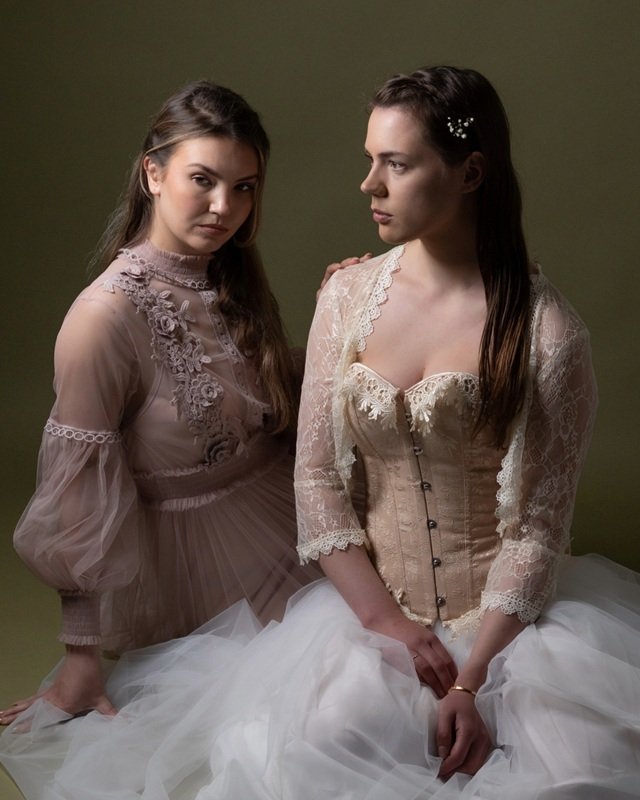 Twee jonge vrouwen in vintage witte en roze jurken, zittend, met een groene achtergrond, het geeft een klassieke romantische sfeer, fine art fotografie.