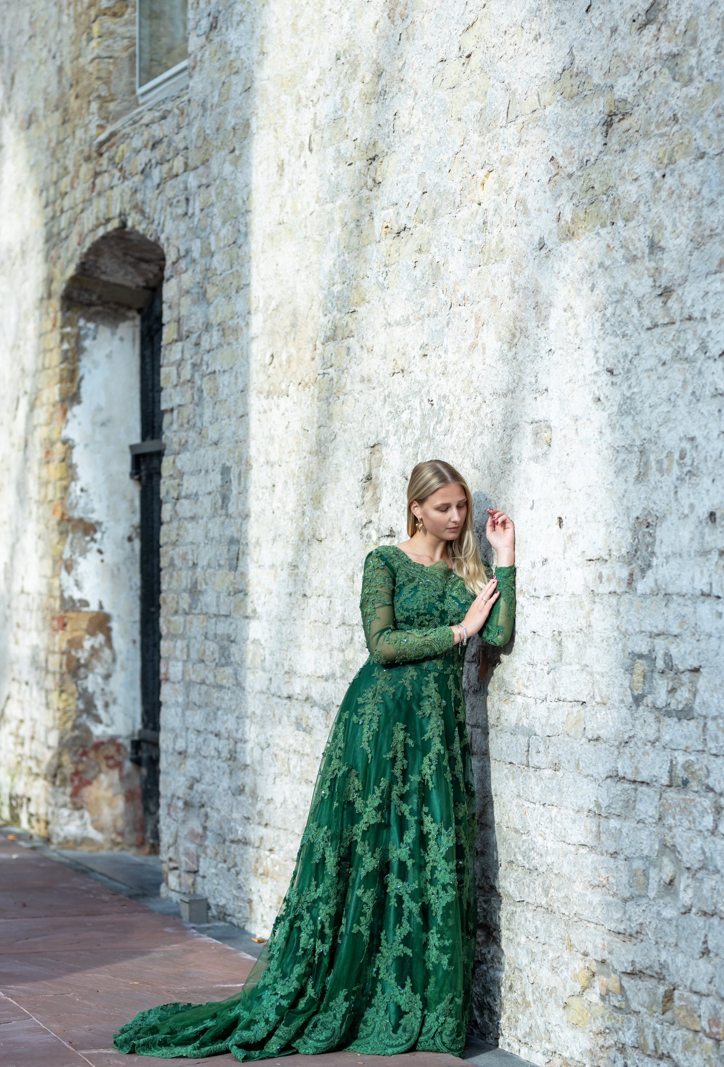 Een blonde vrouw in een groene, elegante avondjurk staat tegen een oude, witte bakstenen muur en kijkt heel dromerig tijdens de fotosessie, daglicht en schaduw