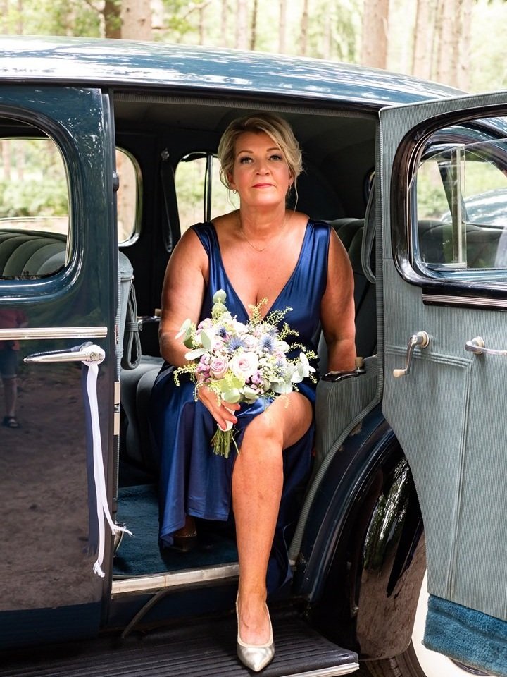Vrouw in een blauwe trouwjurk zit in een vintage auto, houdt een bloemenboeket vast, met een bosrijke achtergrond tijdens een bruiloft in Drenthe