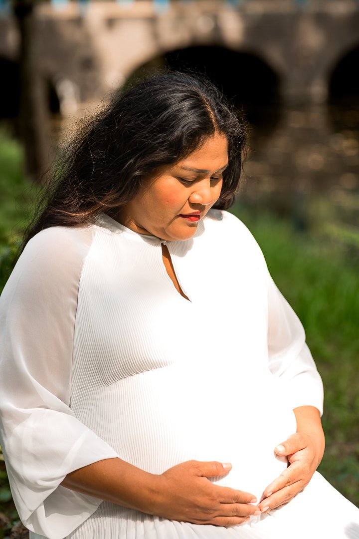 Zwangere vrouw, die heel rustig zit en ondertussen haar buik vasthoudt met haar beide handen, in een groene buitenomgeving, zwangerschapsshoot