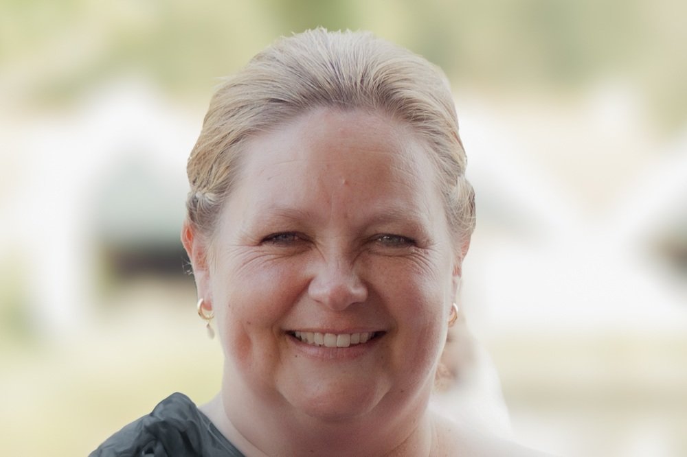 Close-up van een lachende vrouw met blonde haar, buiten op een dag met een groene en lichte achtergrond.