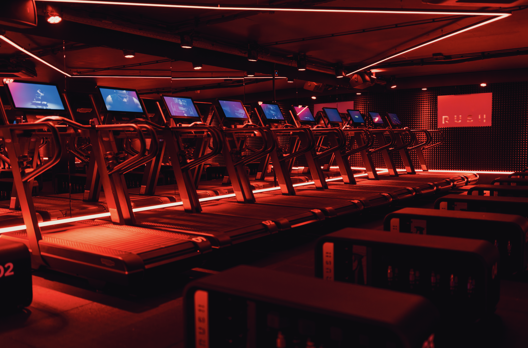 A dimly lit gym with multiple treadmills illuminated by red lighting and digital screens on display.