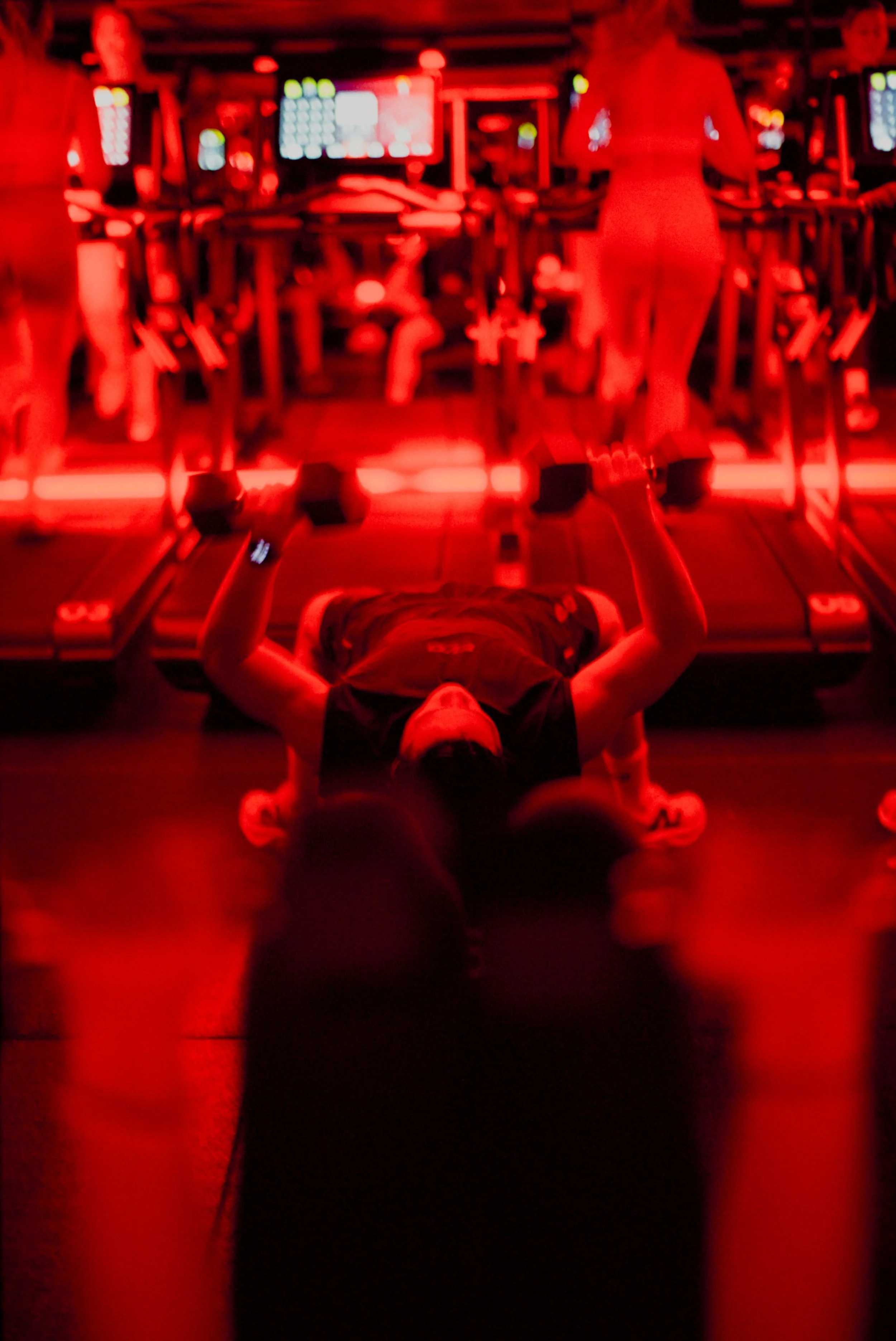 Person lying on the floor of a dimly-lit gym, lifting dumbbells, with treadmills and other gym equipment in the background illuminated by red lights.
