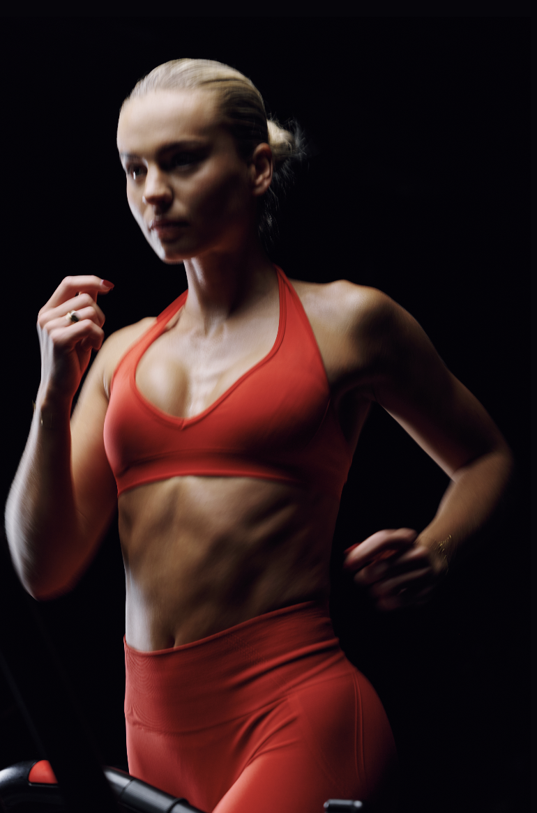 A woman in a red sports bra and matching leggings running on a treadmill in a dark room.
