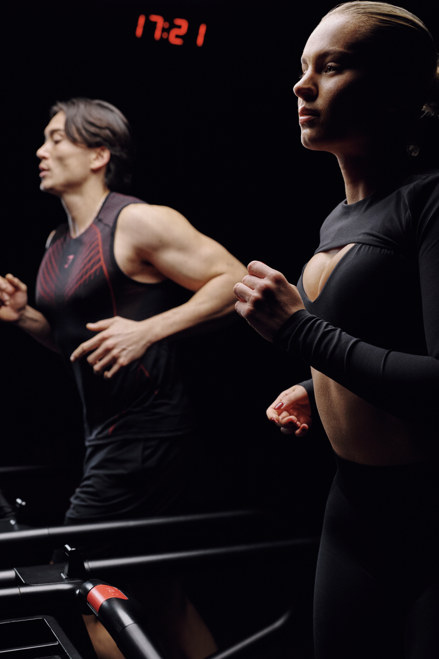 A man and woman running on treadmills in a dark gym, with a red digital clock displaying 17:21 above them.