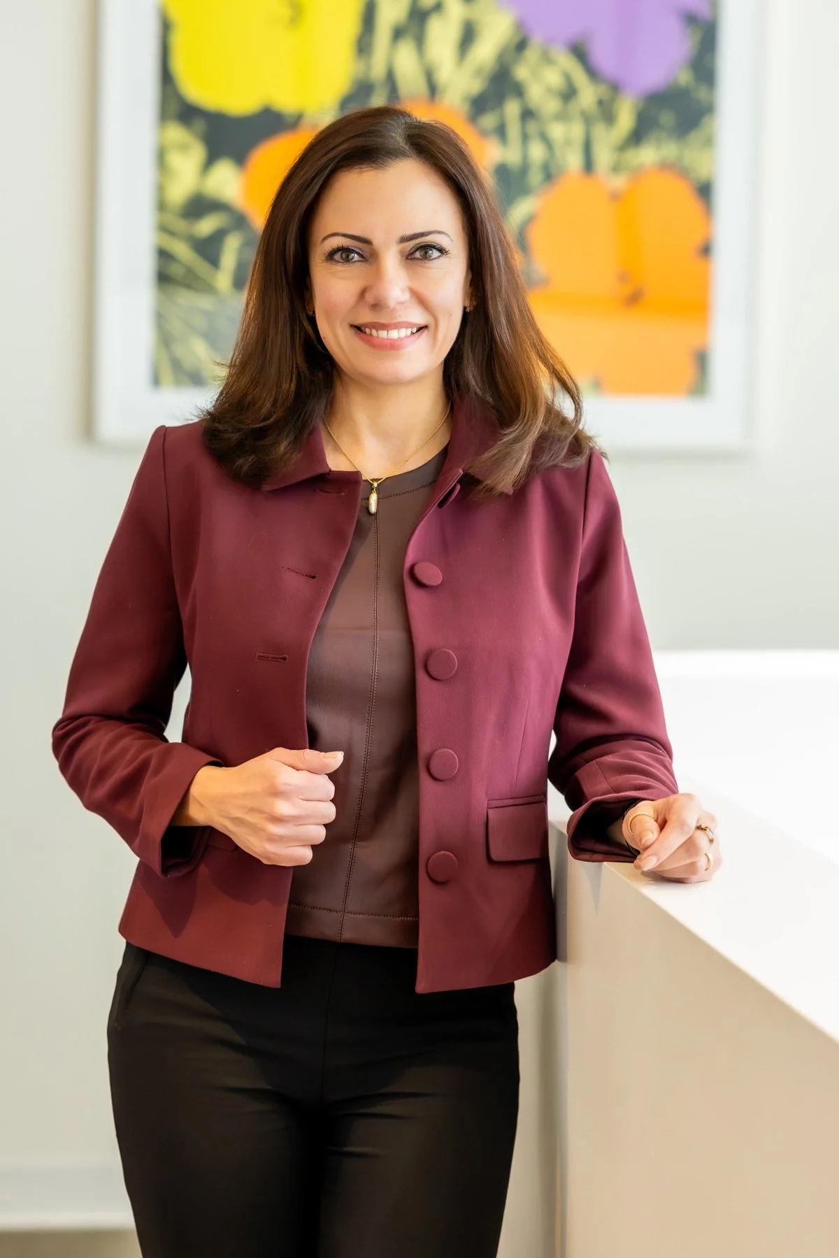 A woman with shoulder-length brown hair, wearing a maroon blazer and dark pants, smiling indoors in front of a colorful abstract painting.