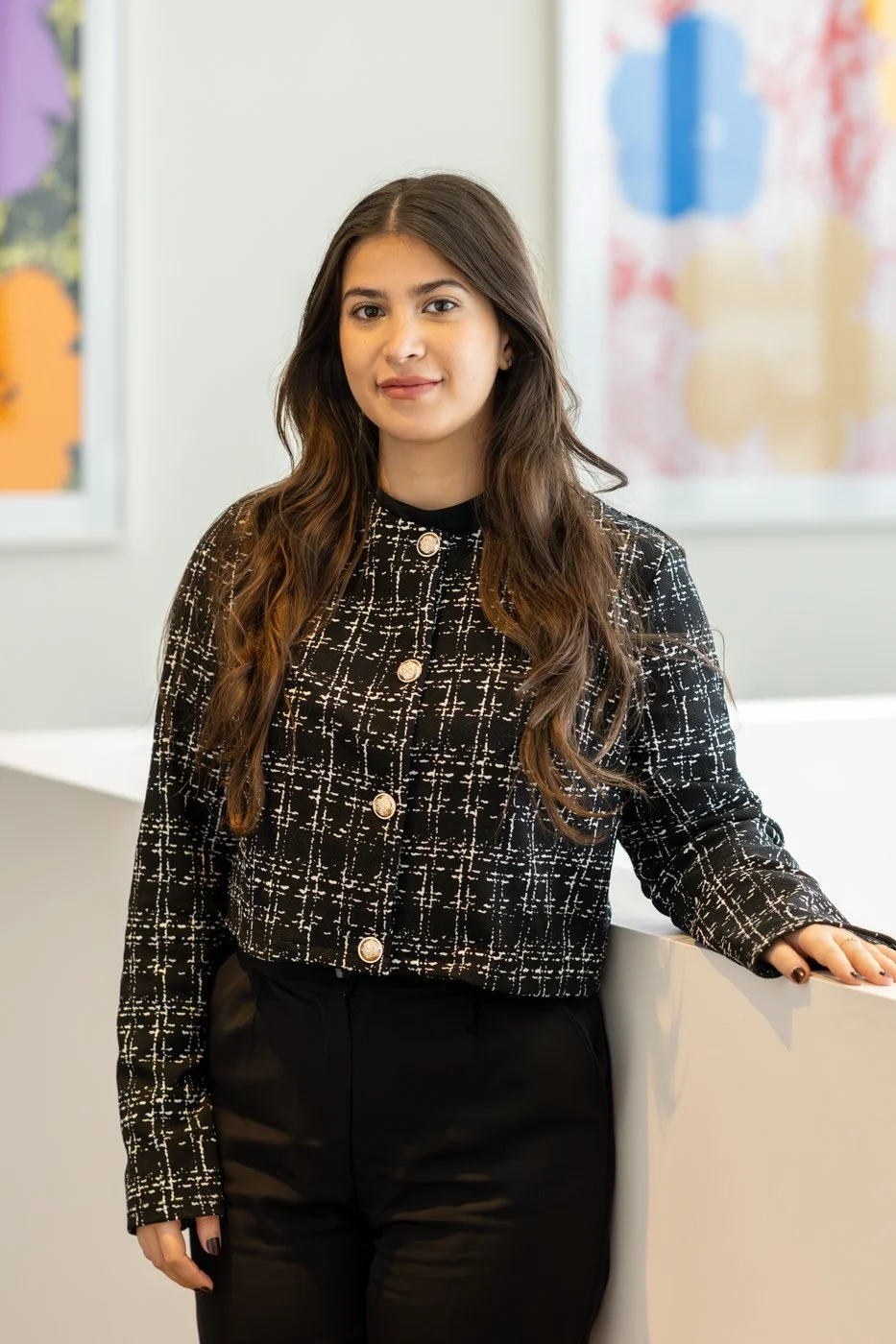 A young woman with long brown hair, wearing a black and white patterned jacket and black pants, stands indoors near a white counter with colorful abstract art in the background.