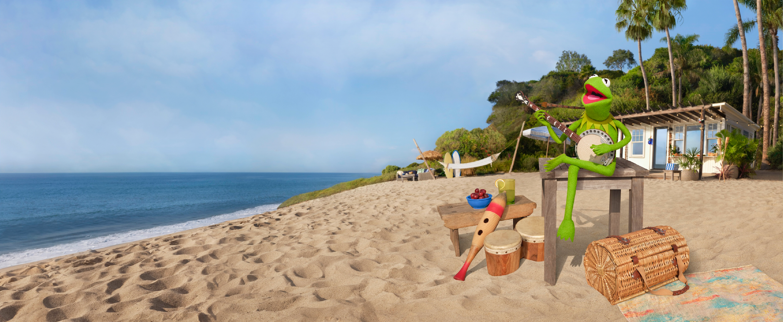 Kermit the Frog sitting on a table playing a banjo on a sunny beach with a house, trees, and lounge chairs in the background.