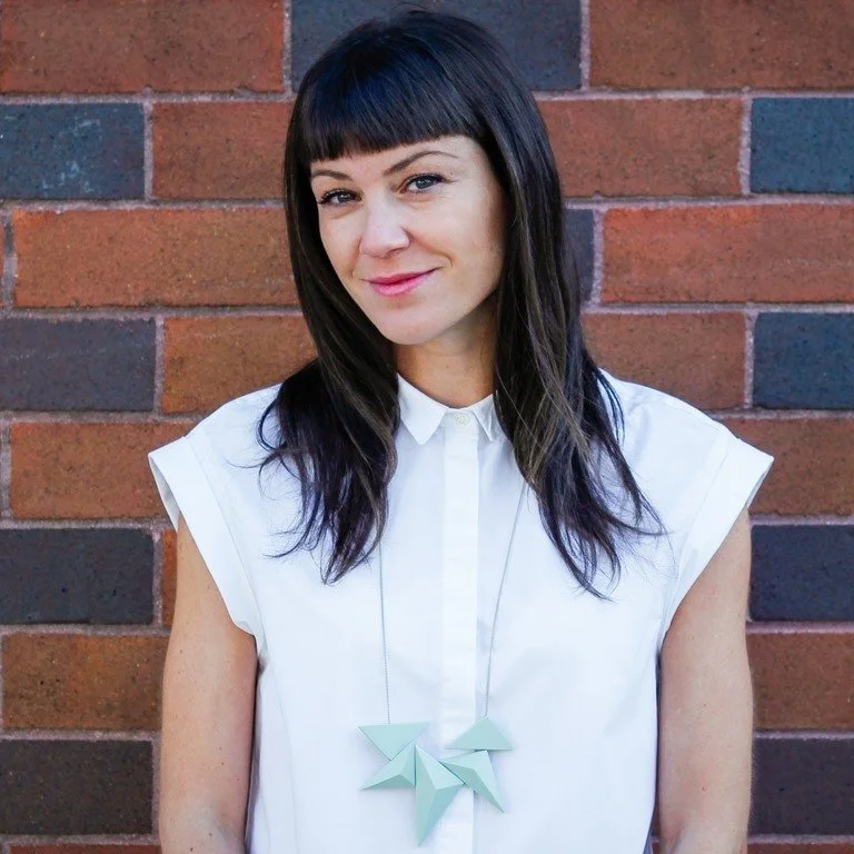 A woman with dark hair and bangs, wearing a white shirt with rolled-up sleeves and a geometric-shaped necklace, standing against a brick wall.