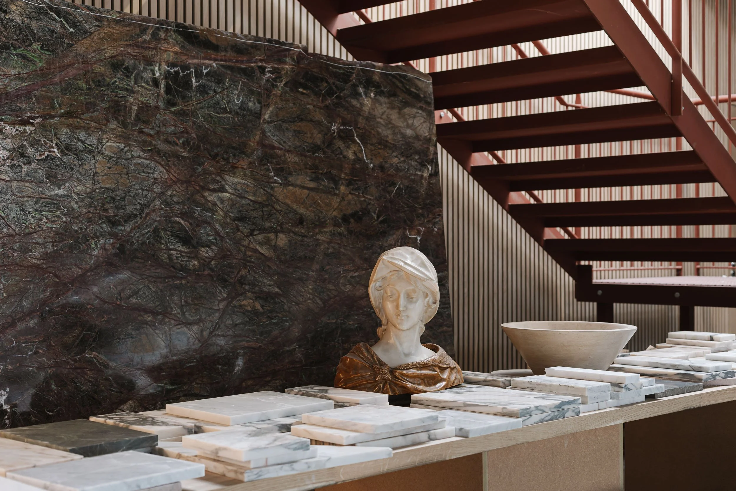 Interior space with a marble sculpture of a woman's head, a large marble bowl, and an array of marble slabs on a wooden table. A dark, veined marble wall and a staircase with reddish-brown steps and a striped wall are visible in the background.