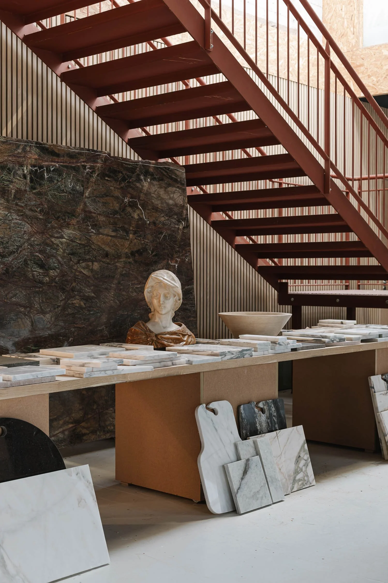 A marble bust of a woman with short hair and a scarf around her neck, placed on a display table among various marble slabs in a showroom, with a decorative staircase overhead and wall paneling in the background.