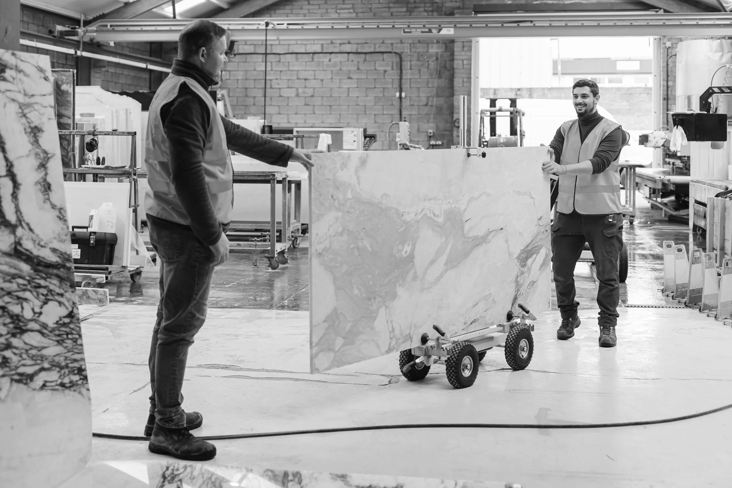 Two workers in a factory handling a large slab of marble or similar stone. One person is guiding the stone on a wheeled cart, while the other observes.