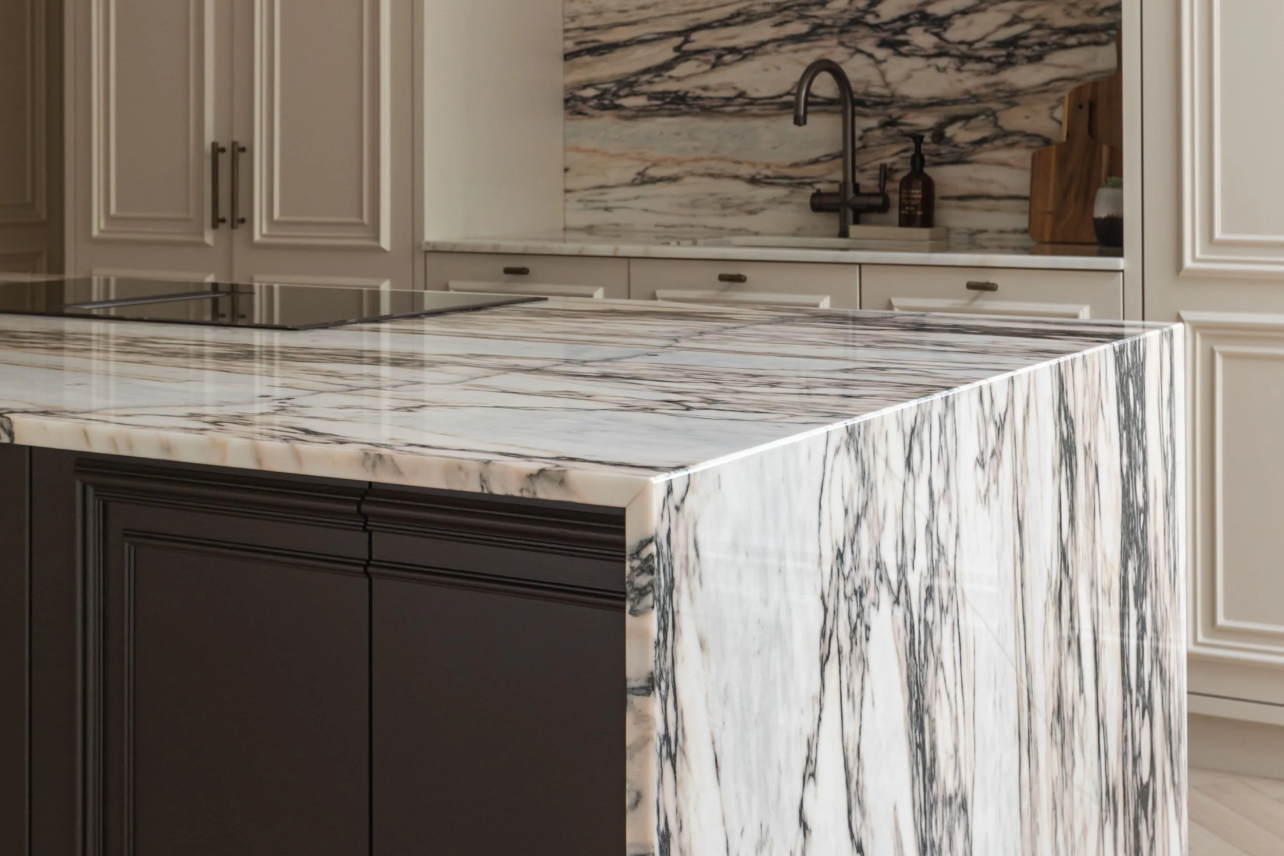 A kitchen with marble countertops and backsplash, cream-colored cabinets, a black faucet, a wooden cutting board, and a bottle of hand soap.