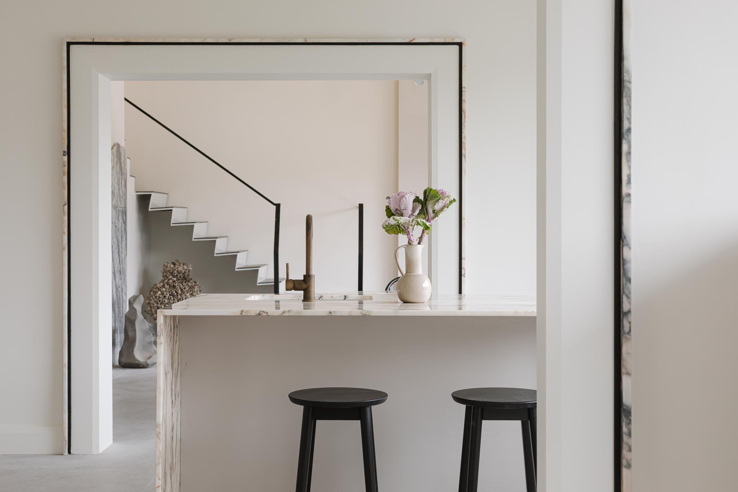 Modern kitchen with a marble breakfast bar, two black stools, a white vase with pink and green flowers, and a view of stairs in the background.