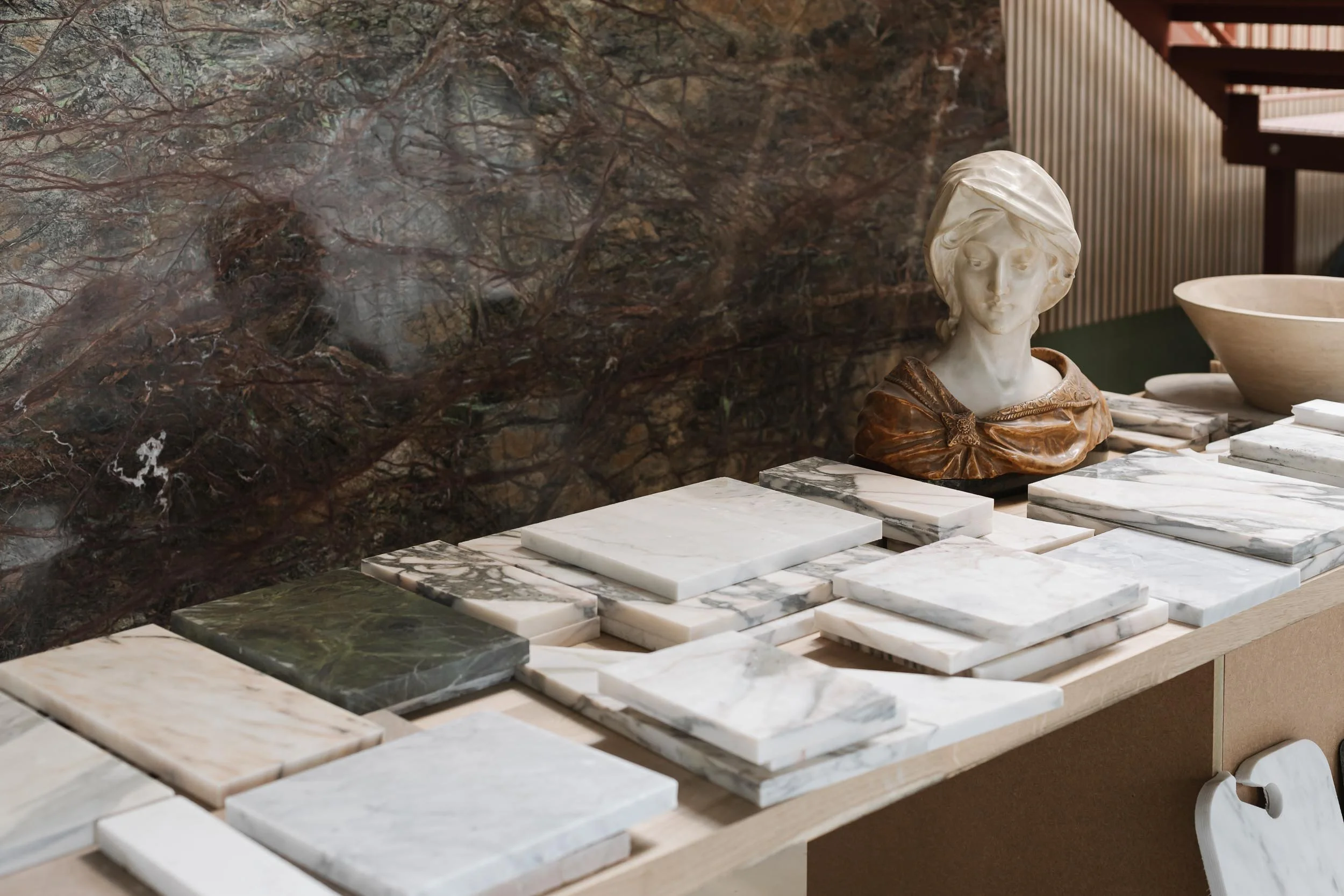 Display of various marble slabs and a bust sculpture of a woman on a marble table, against a dark, textured wall.