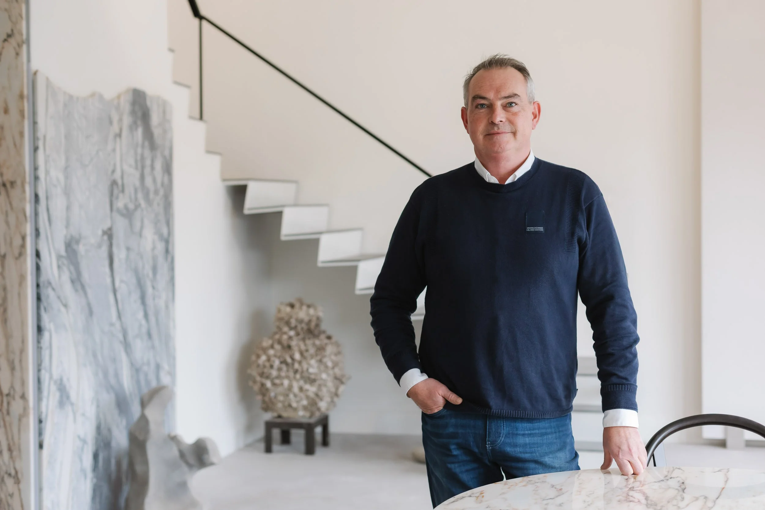 A middle-aged man with gray hair and a slight smile, wearing a dark sweater over a white collared shirt, standing in a modern, well-lit interior with a staircase and decorative art piece in the background.