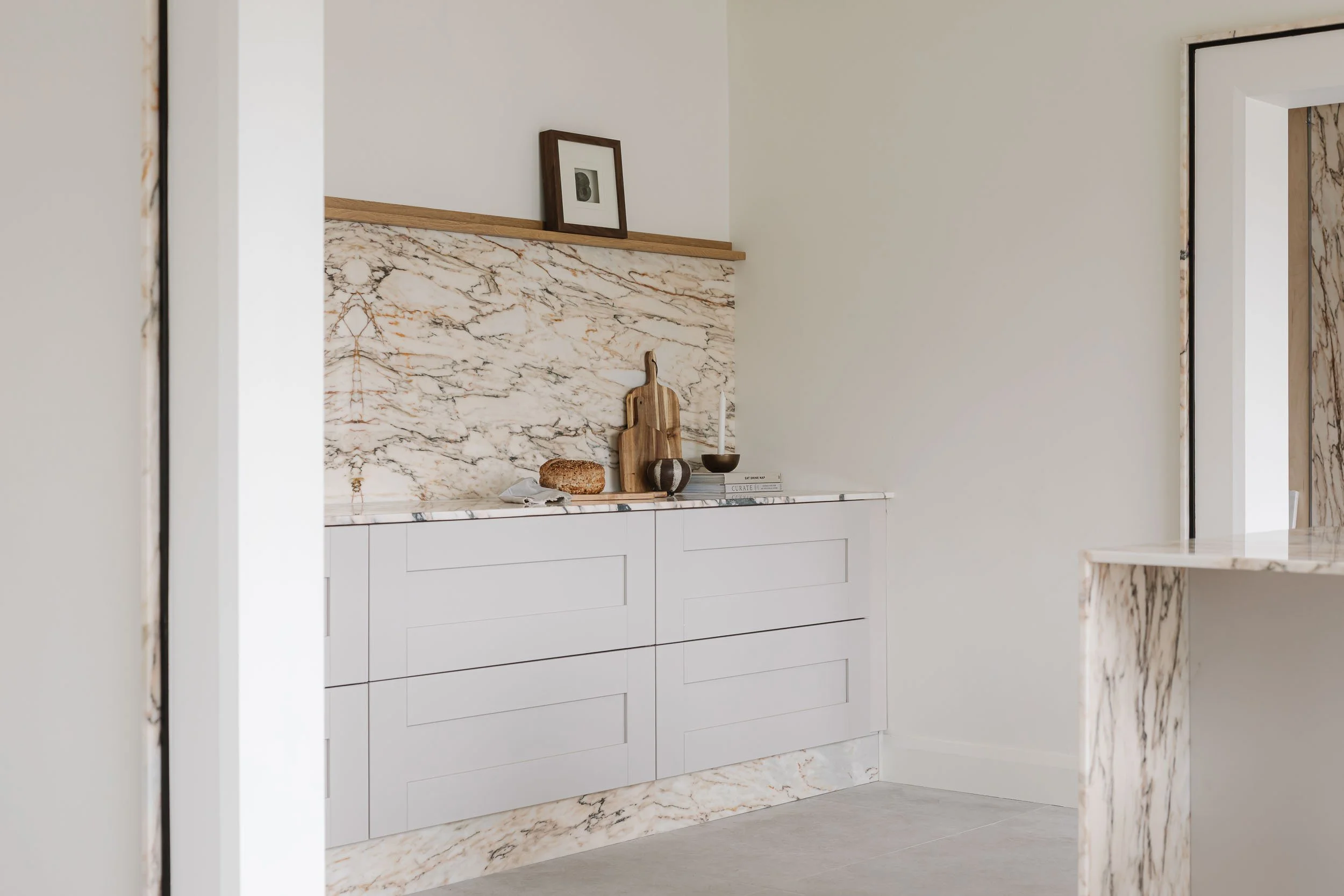 A kitchen with white cabinetry, marble backsplash and countertop, decorated with a cutting board, candle, and a few books, and framed artwork on a ledge.