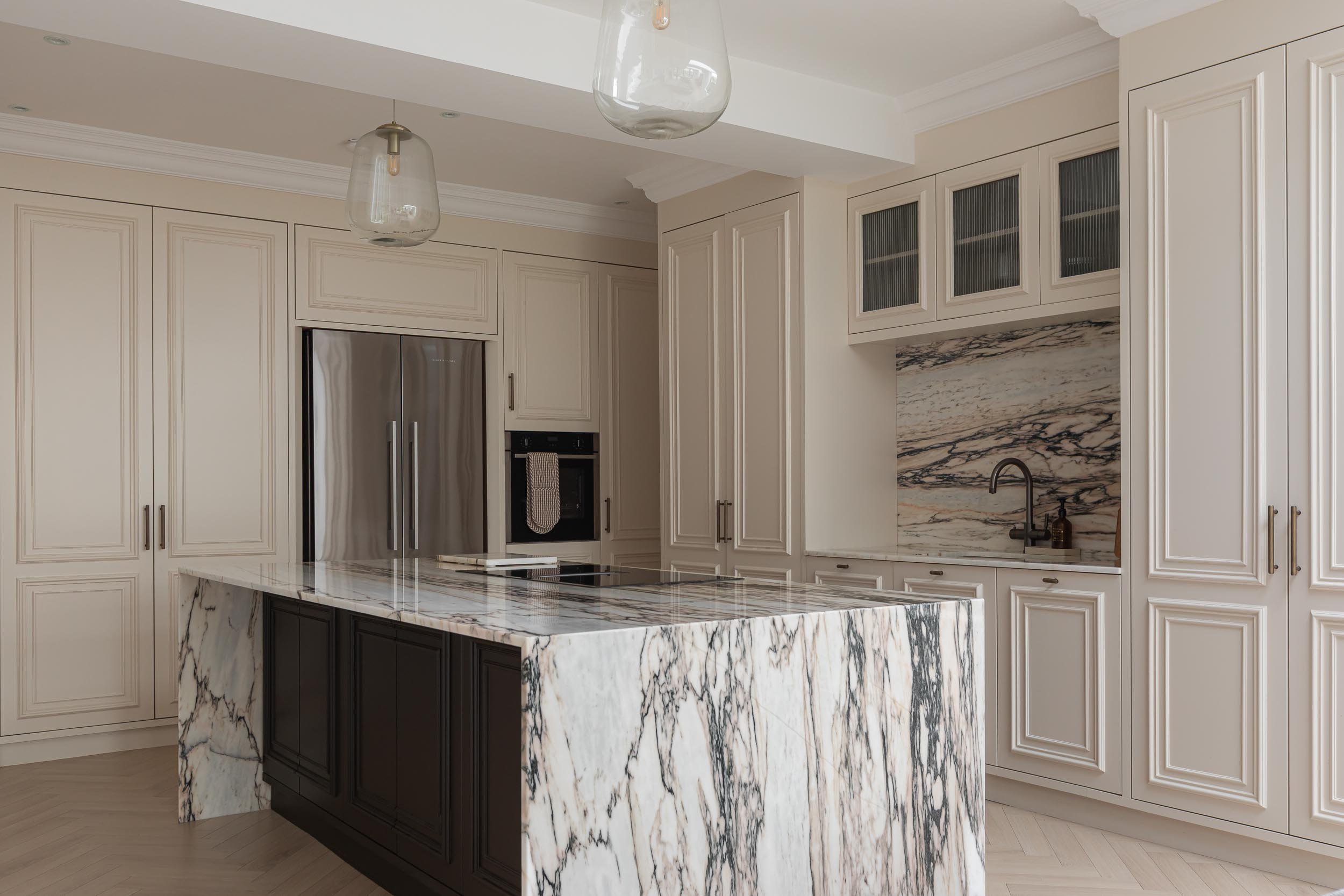 A modern kitchen with white cabinets, a marble island, and a marble backsplash. There are two pendant lights hanging from the ceiling, and a stainless steel refrigerator is visible in the background.