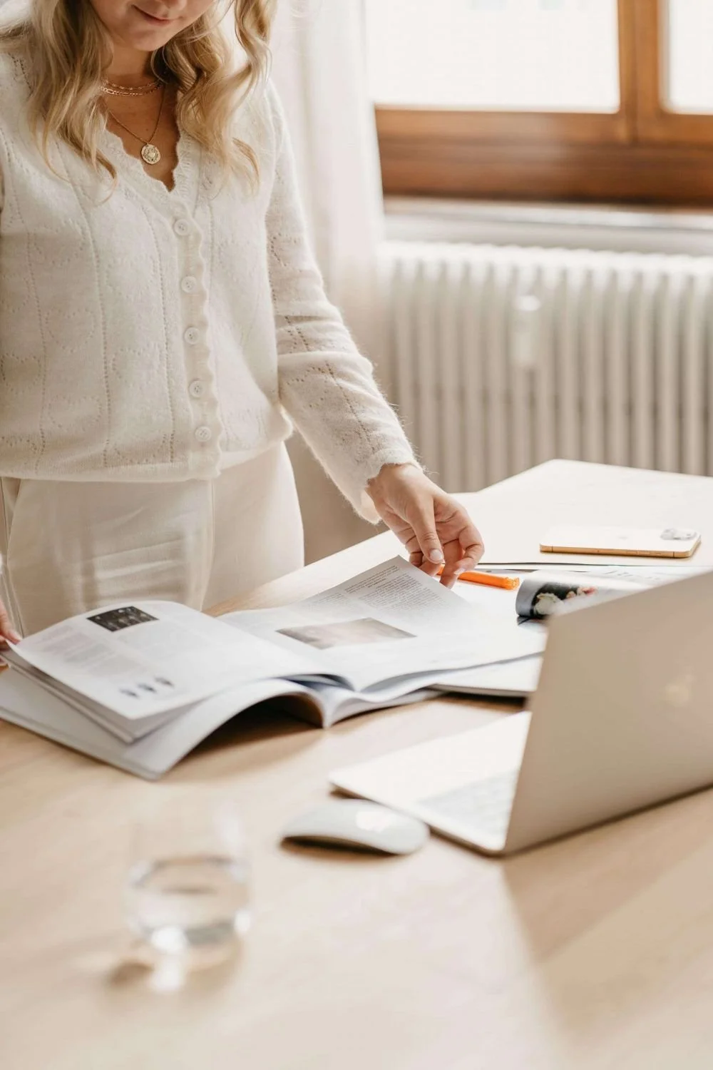 A woman in a cream-colored sweater and jewelry is working at a wooden table, flipping through an open magazine with a laptop, notebook, pen, and glass of water nearby in a bright room with a radiator and large window.