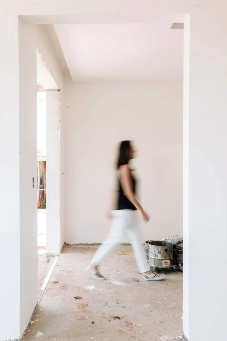A woman walking inside an unfinished room with construction equipment on the floor.