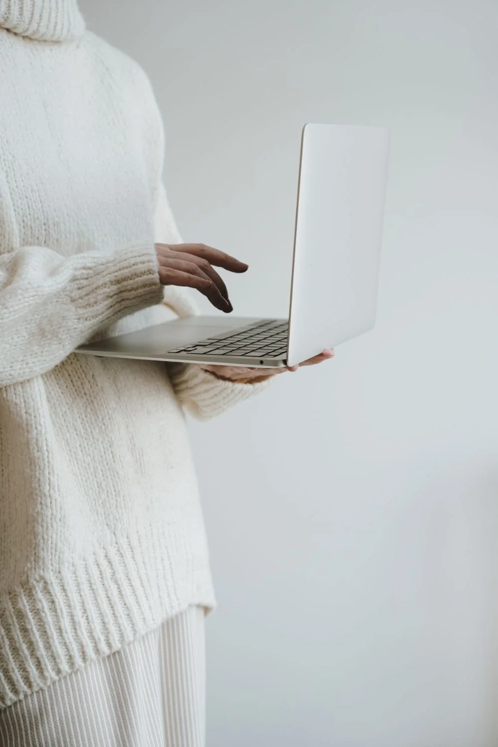 Person in a cream, knitted sweater holding an open silver laptop against a plain light grey background.