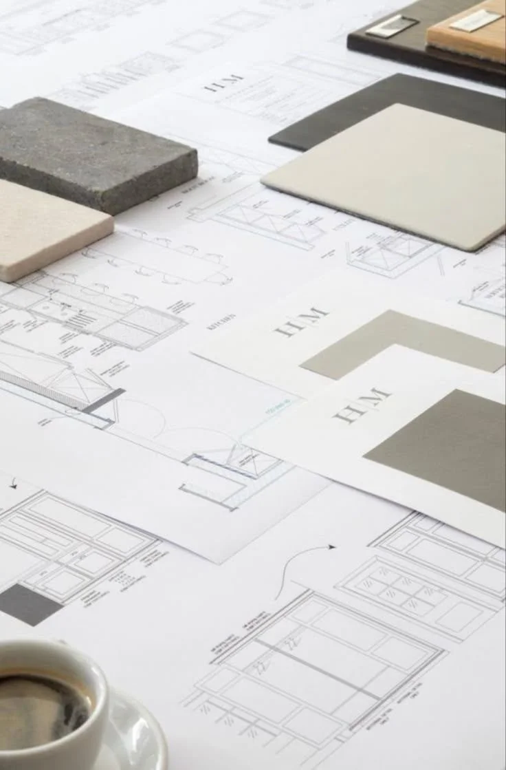 Architectural blueprints and sample materials on a workspace table, with a cup of coffee in the foreground.