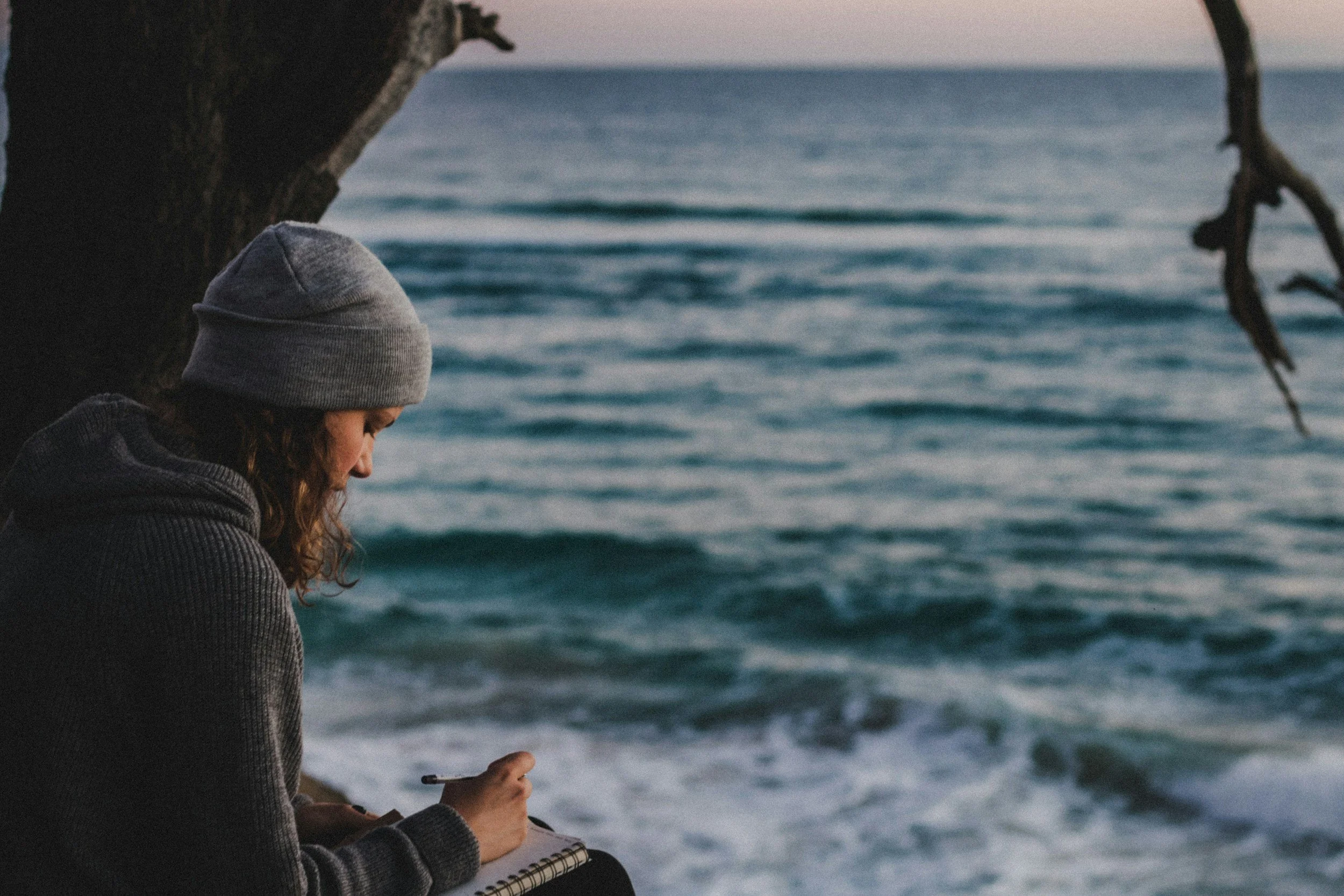 A person wearing a gray beanie and dark jacket writing or drawing in a notebook while sitting under a tree near the ocean during sunset.