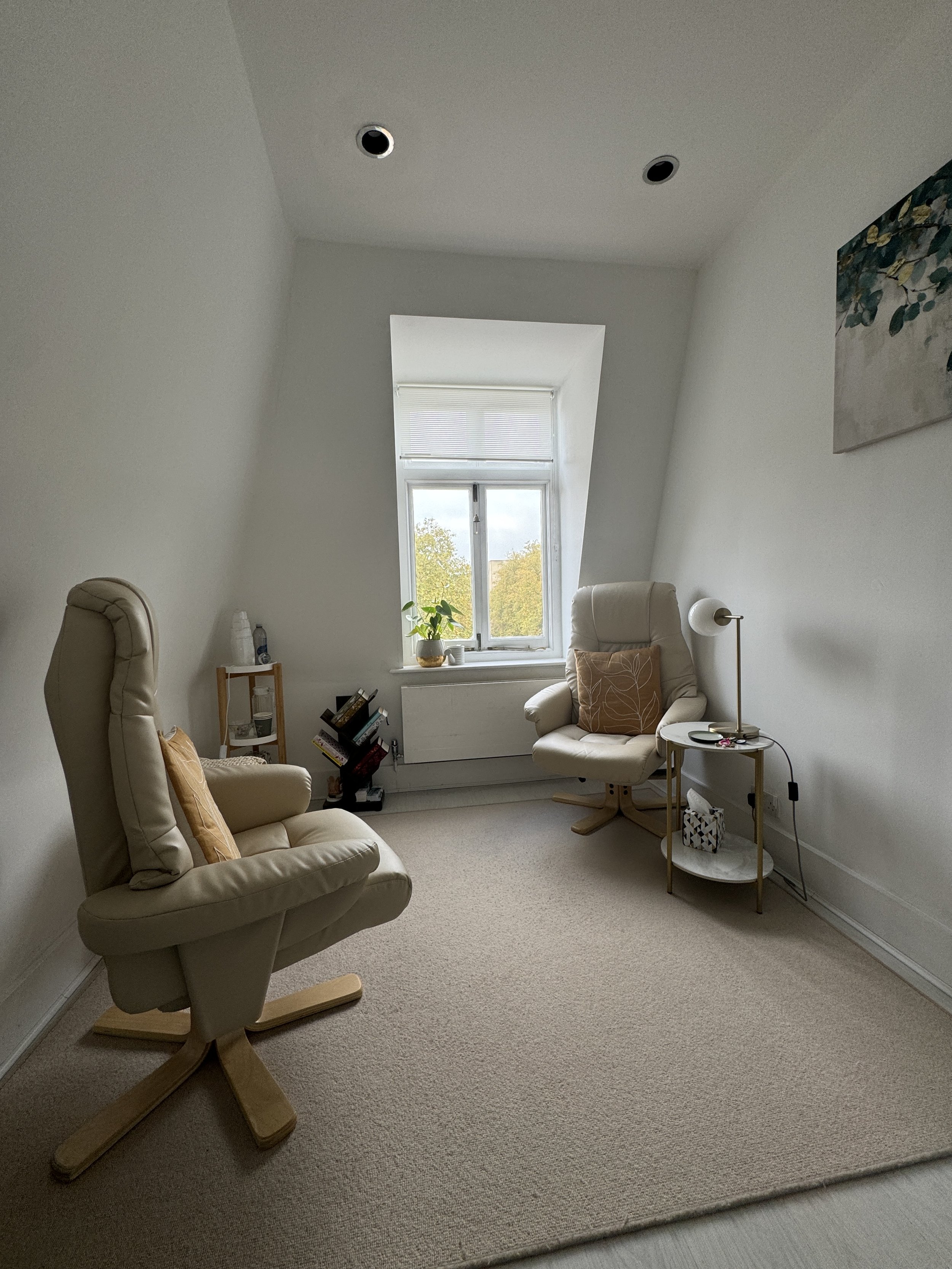 A cozy reading nook with two off-white armchairs, a small side table, a window with a view of trees, a potted plant, and wall art, in a bright, minimalist room.