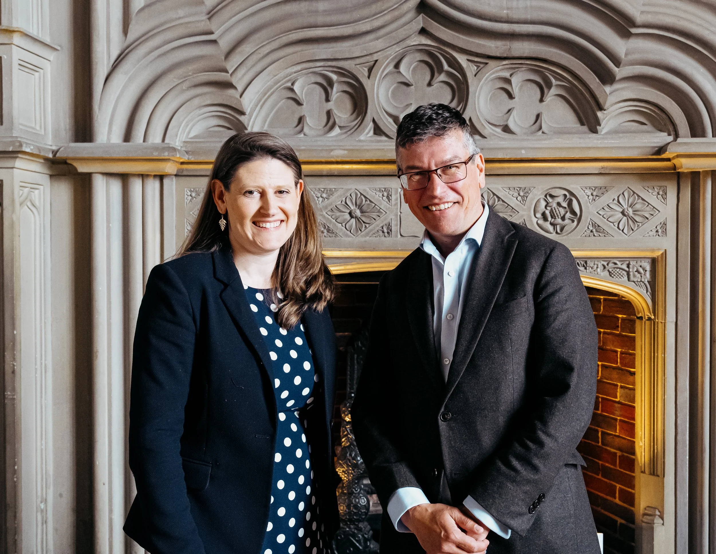 A woman and a man smiling and standing close together in front of an ornate fireplace with carved designs and a gold trim in a formal setting.