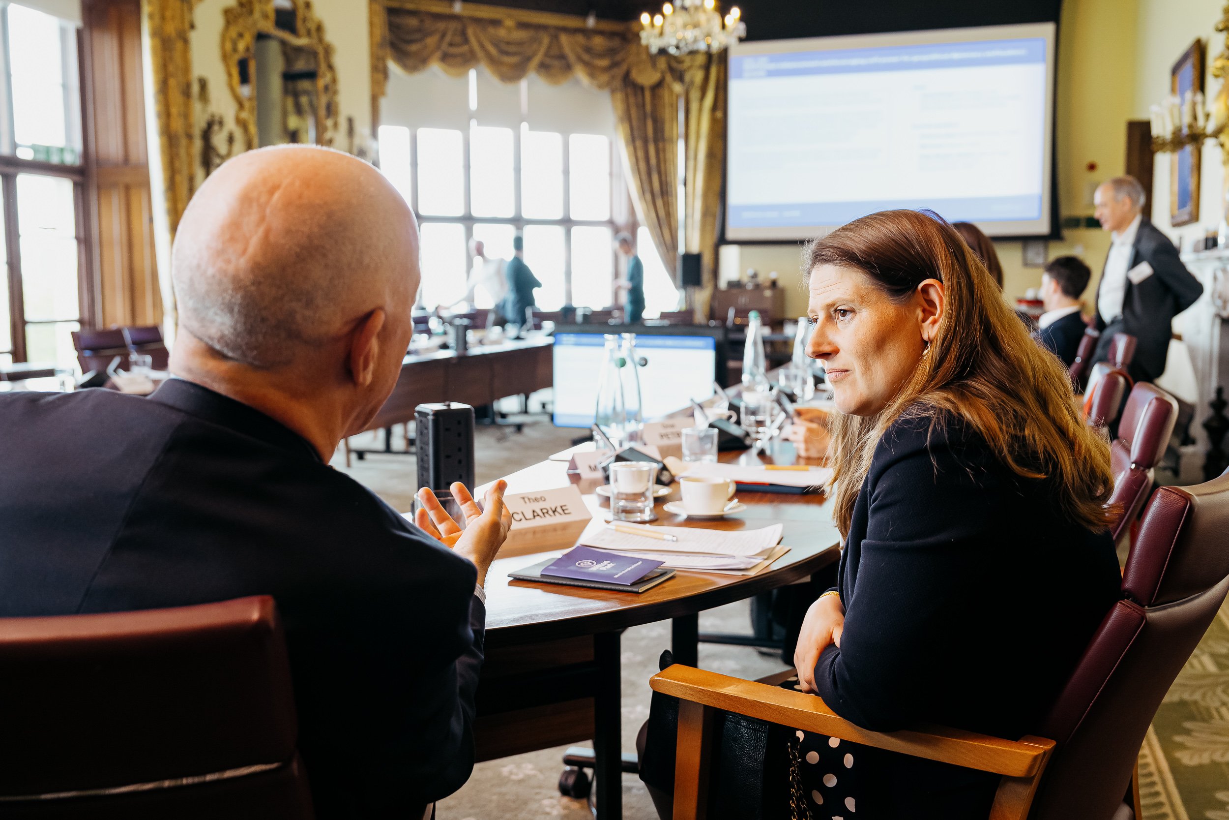 Two women are sitting at a conference table engaged in conversation during a meeting in a well-lit room with large windows, chandeliers, and a large screen in the background. Other people are present in the room, some standing and talking.