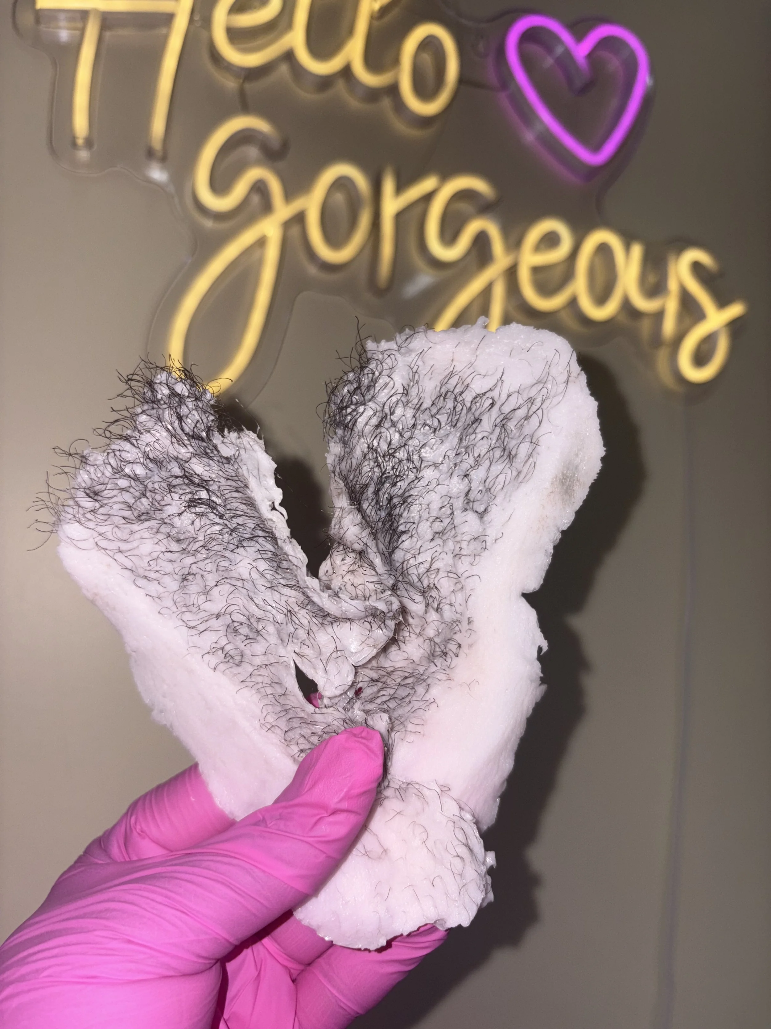 A hand wearing a pink glove holding a piece of cotton with some hair attached, against a background with a neon sign that says 'Hello Gorgeous' and a purple heart.