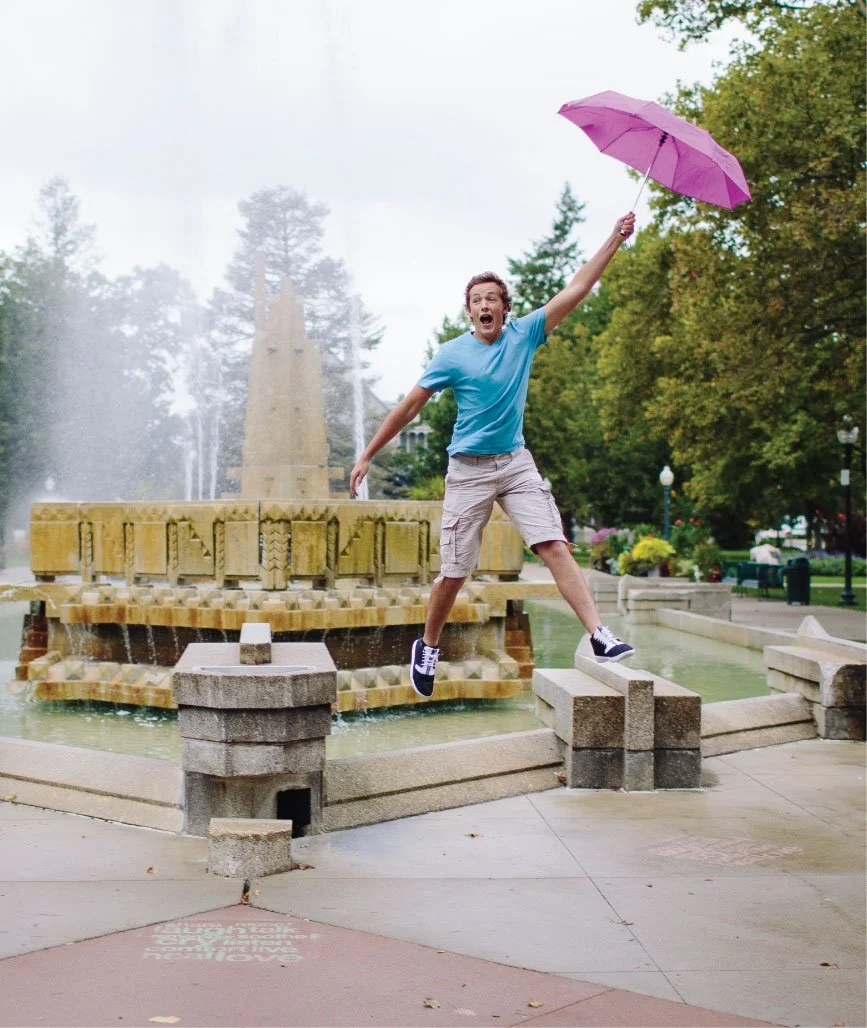 A young man jumping in front of a fountain in a park, holding a purple umbrella, surrounded by green trees and park benches.