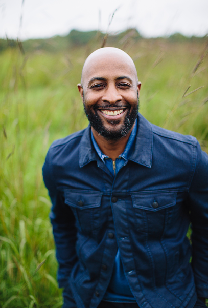 A smiling man outdoors in a grassy field, wearing a dark blue jacket.