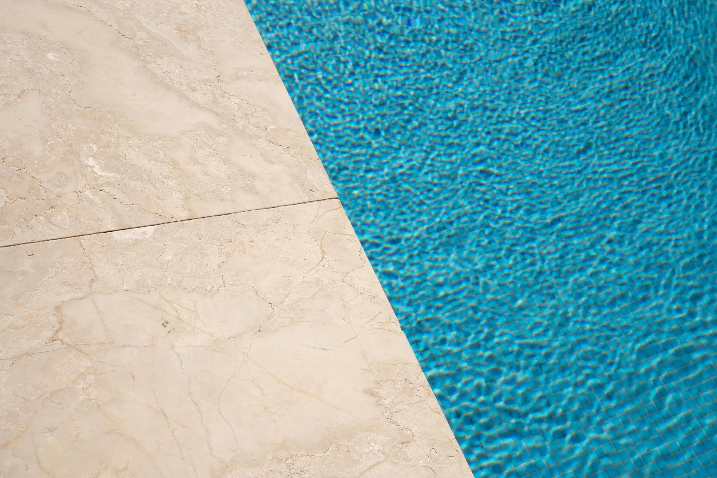 Close-up of a swimming pool with blue water and beige stone tile surrounding it.