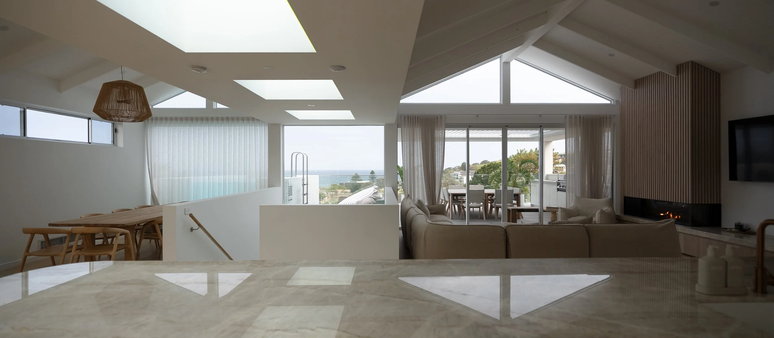 Modern living room with large windows, sliding glass doors leading to a balcony, ocean view, beige sectional sofa, fireplace, and dining area with wooden table and chairs.