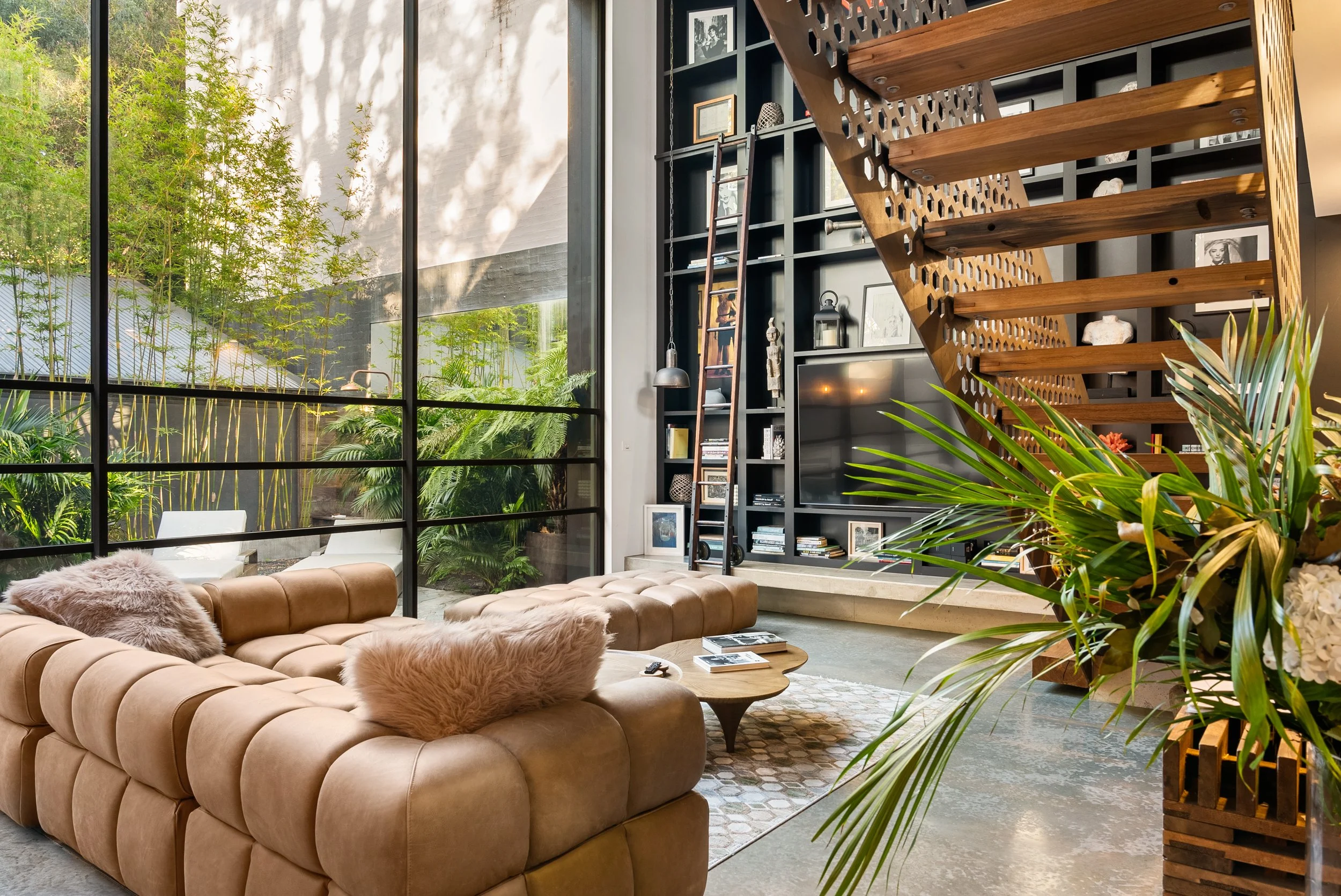 Modern living room with large floor-to-ceiling window showing lush outdoor greenery, tan tufted leather sofa with furry pillows, black built-in bookshelf with decor, ladder, and flat-screen TV, wooden staircase with open risers, potted plants, and contemporary decor.