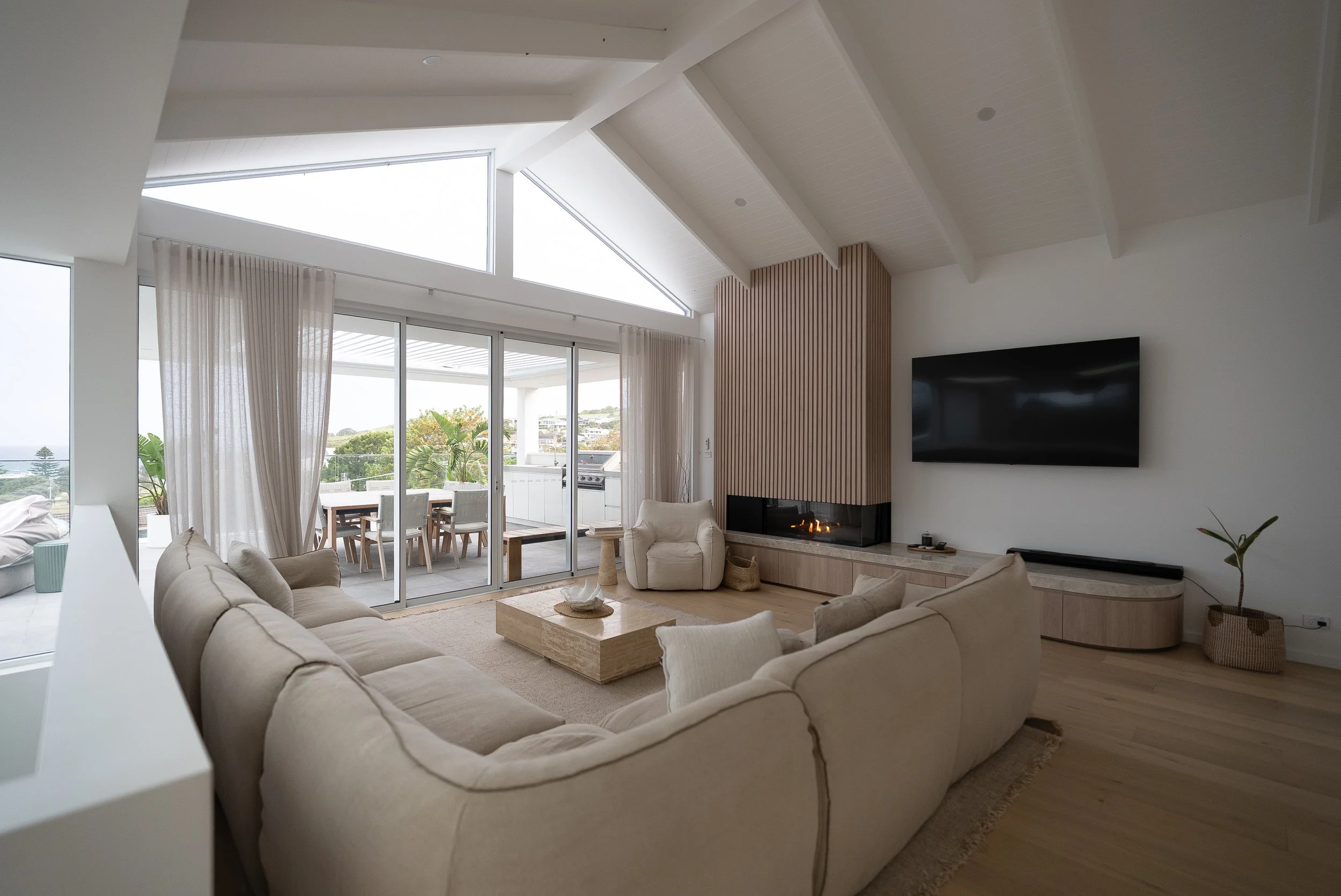 Living room with a large beige sectional sofa, armchair, fireplace, flat-screen TV, sliding glass doors opening to an outdoor patio with a dining table, and a bright, airy interior with high vaulted ceilings and white walls.